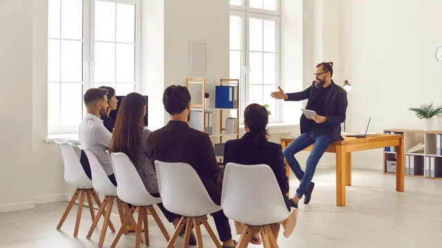 Formador sentado en una mesa habla con un grupo de profesionales en una sala luminosa.